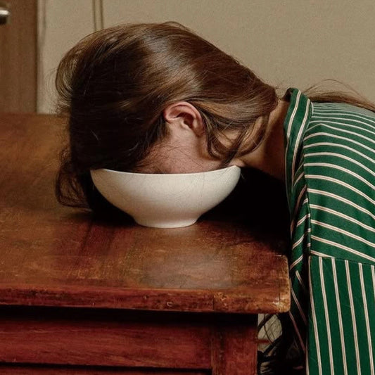 A woman resting her face in a white bowl on a wooden table